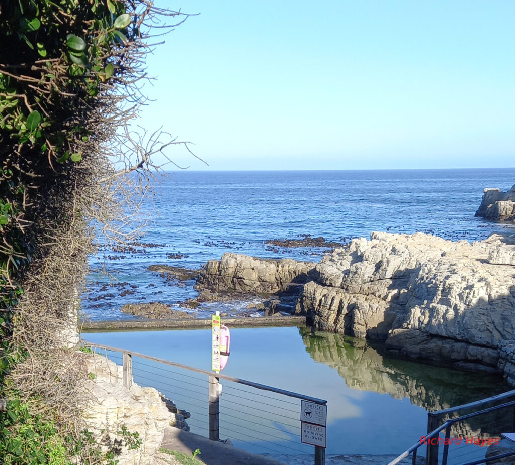 A small tidal pool surrounded by rocks and a single low wall facing out to sea.
Kelp can be seen in the sea beyond the wall.