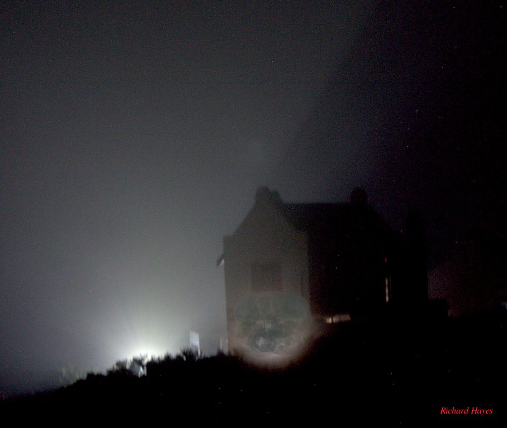 Car headlights illuminate the mist and the side of a house in the Cape Dutch Architectural style.